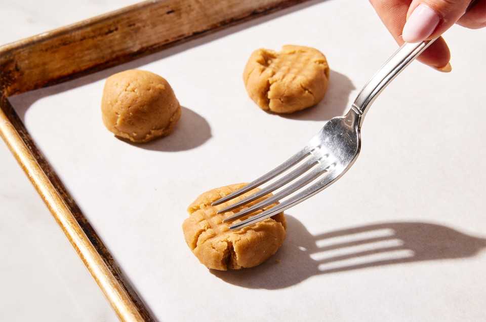 A baker pressing scooped balls of peanut butter cookie dough with the back of a fork on a baking sheet - select to zoom