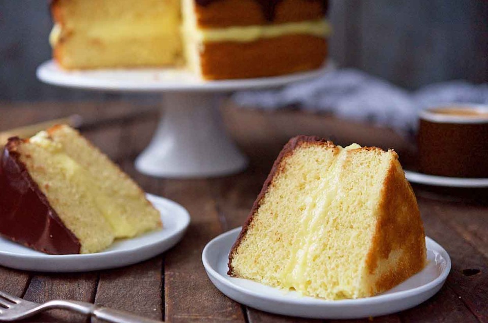 A Boston Cream Pie on a cake stand featuring two layers of yellow cake filled with pastry cream and a chocolate ganache, with two slices on plates in front of it. - select to zoom
