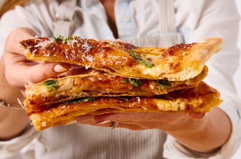 A person holding three slices of flaky puff crust pizza and showing the laminated and crispy crust, and the melty cheese.