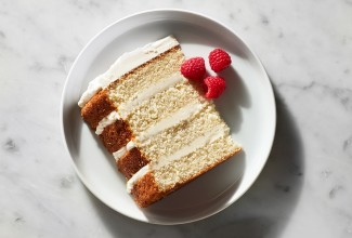 A slice of a four-layer Tender White Cake filled with vanilla buttercream and garnished with raspberries on a plate