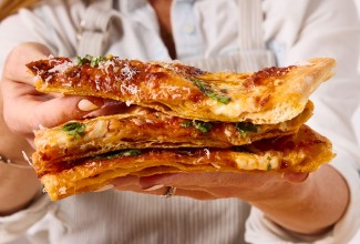 A person holding three slices of flaky puff crust pizza and showing the laminated and crispy crust, and the melty cheese.