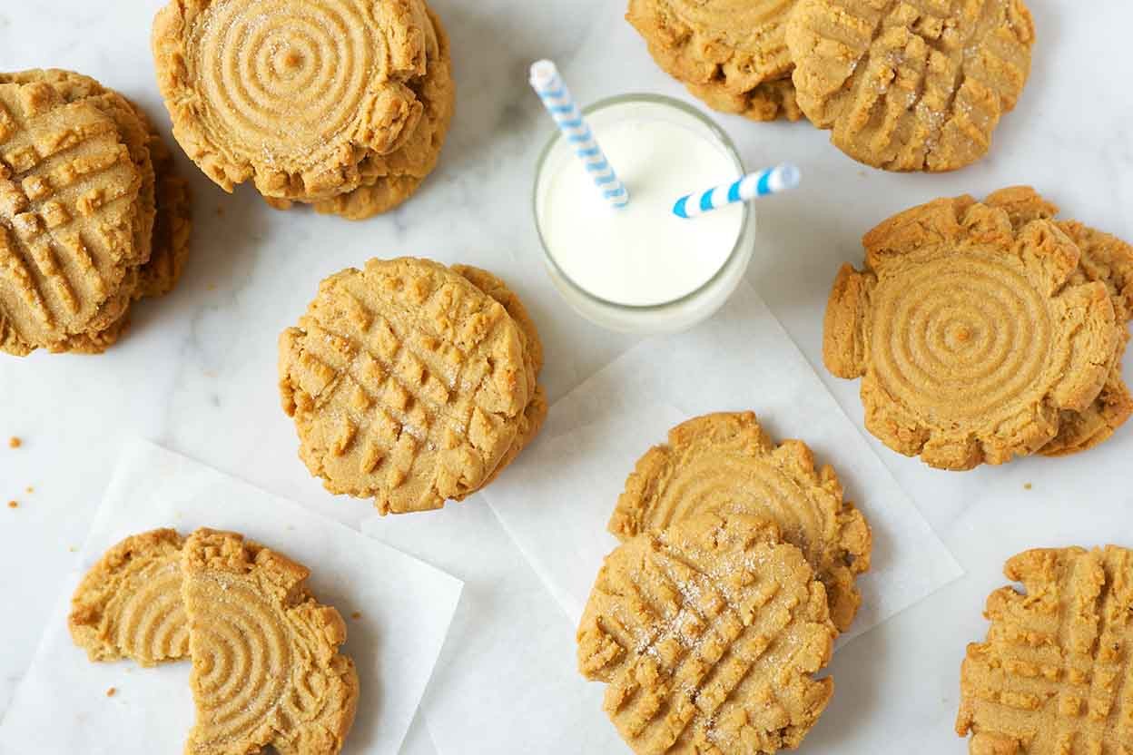 An assortment of Classic Peanut Butter Cookies on a kitchen table next to a glass of milk with a straw